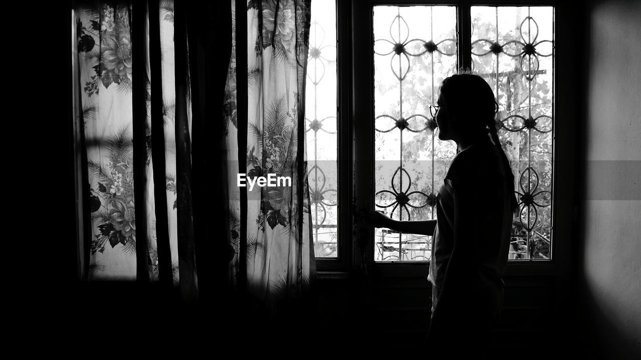 Side view of silhouette teenage girl looking through window at home