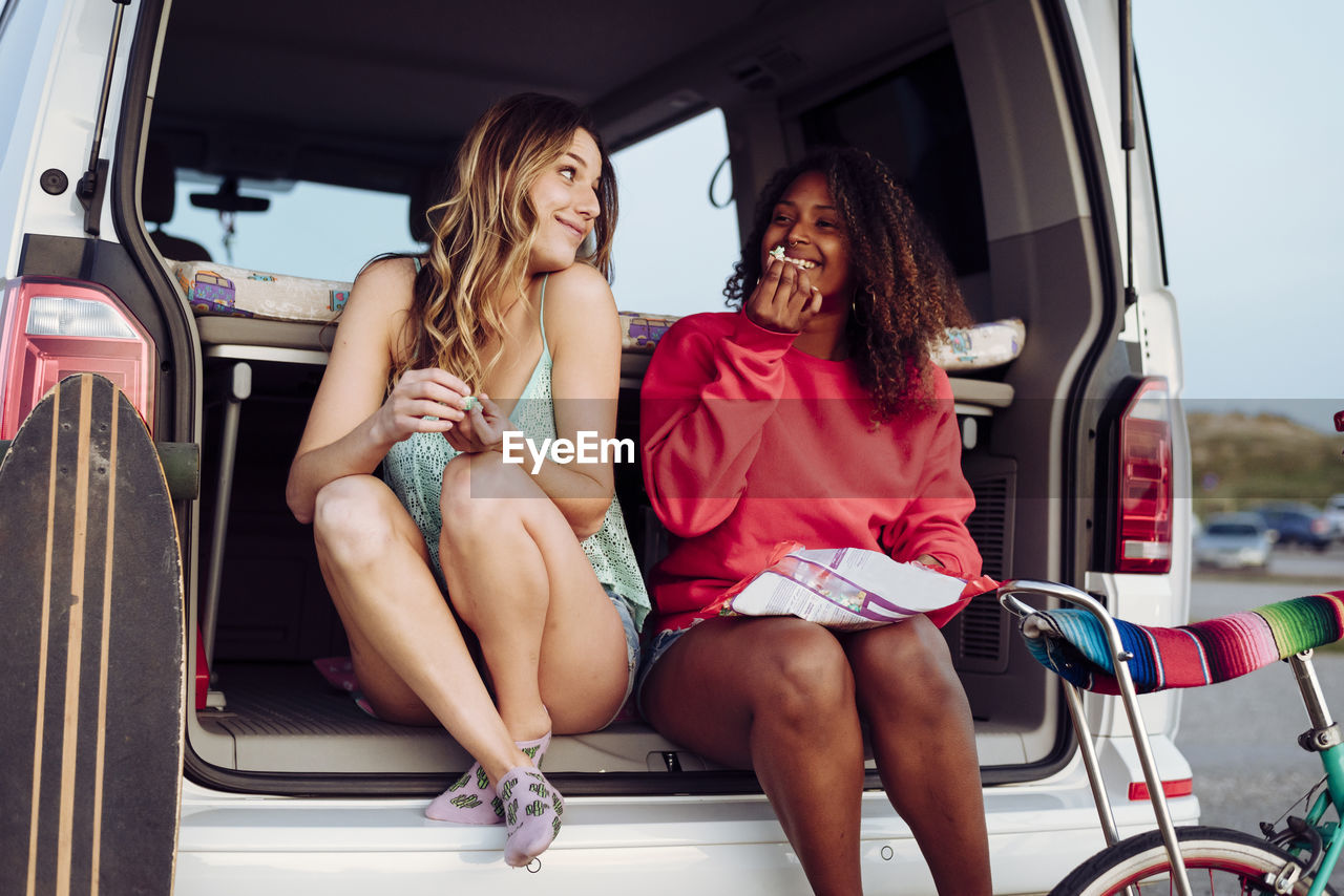 Cheerful women looking at each other while sitting on trunk of van eating popcorn