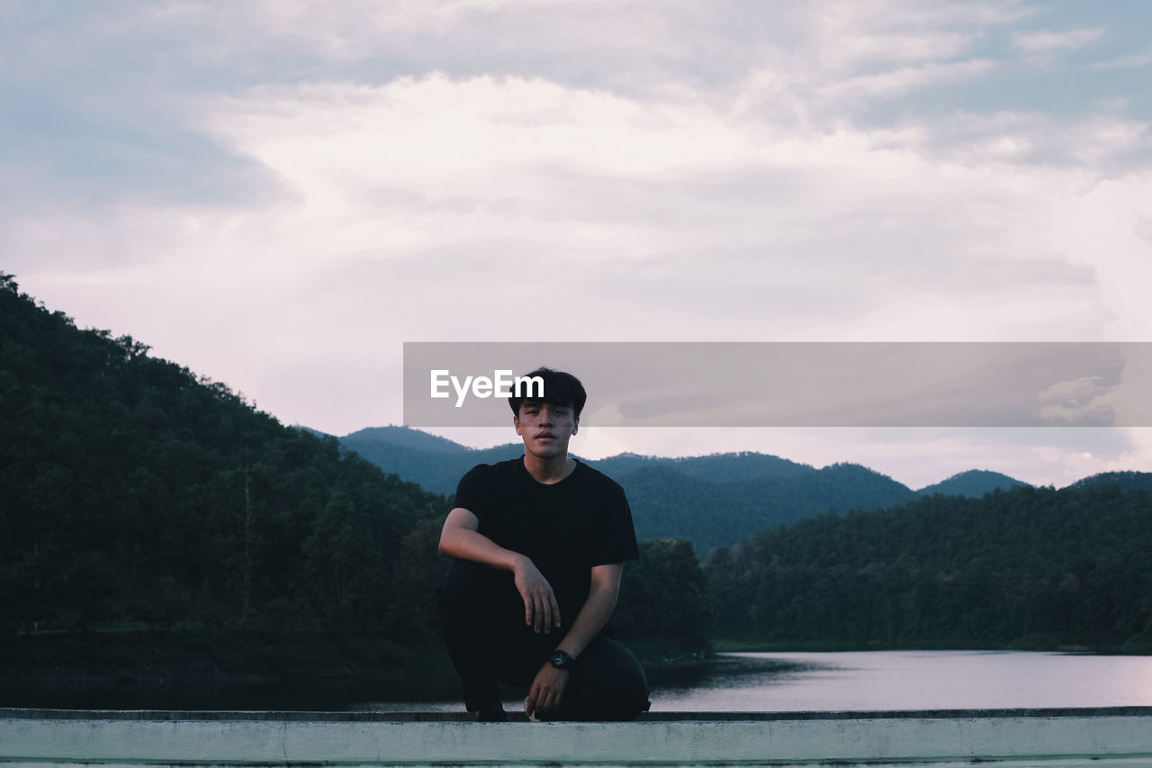 Full length of young man crouching on retaining wall against river and forest