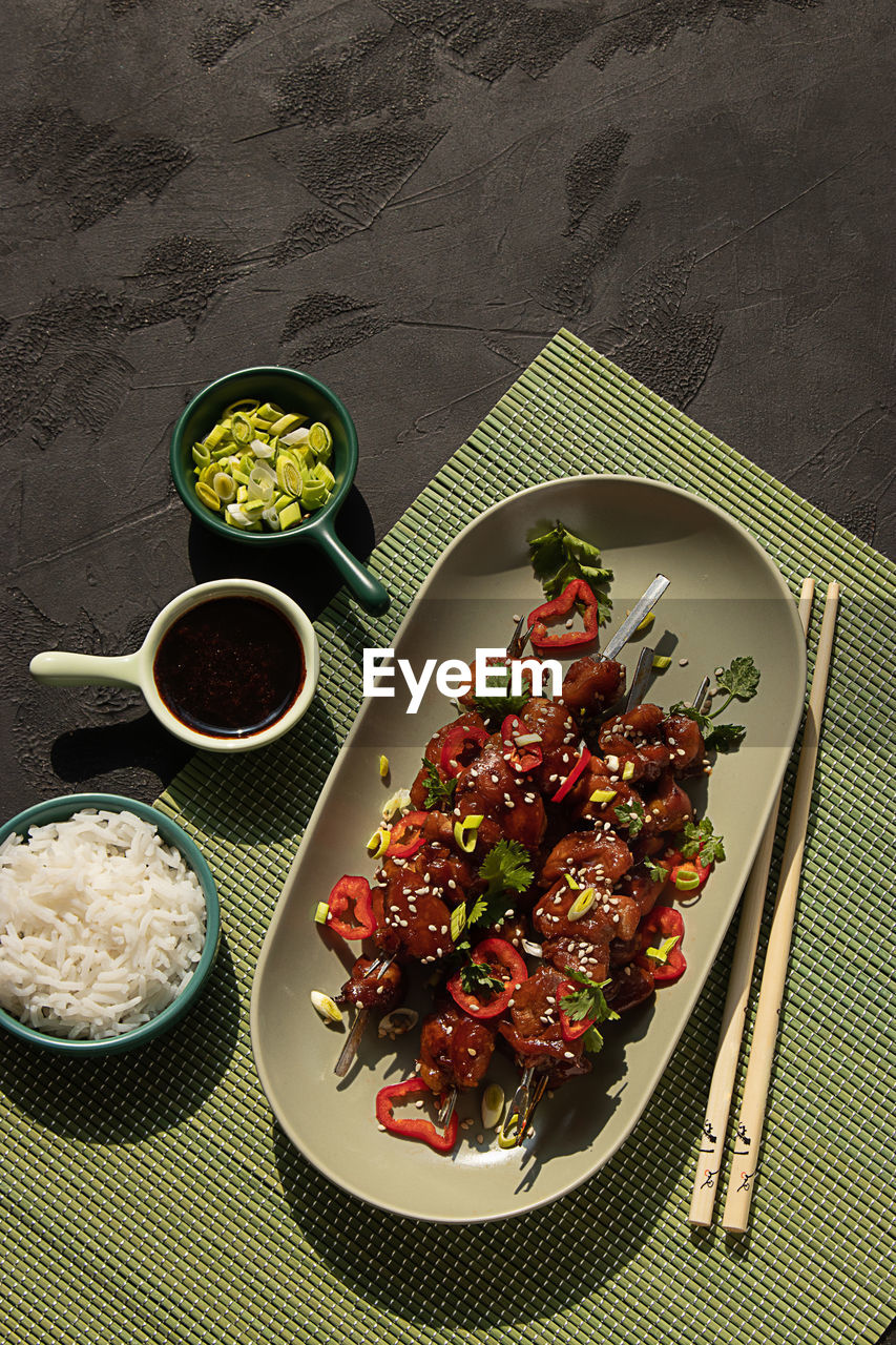 Top view of delicious marinated chicken on skewers topped with greens served on table with bowls of rice, soy sauce and leek in restaurant