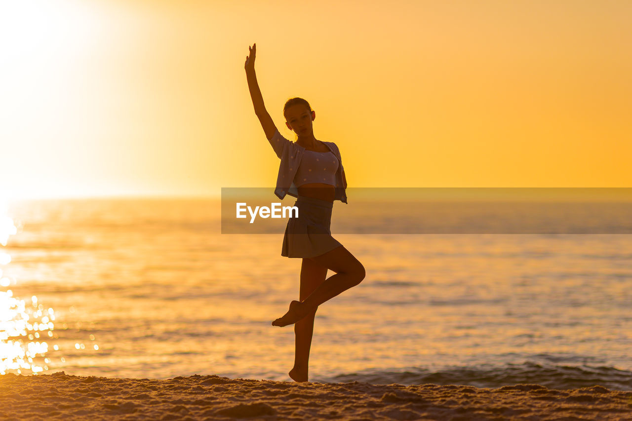 side view of silhouette woman with arms outstretched standing at beach during sunset