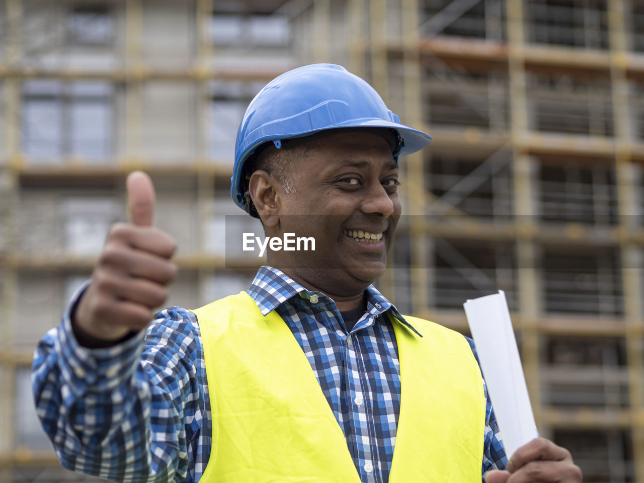 Portrait of a successful indian engineer holding rolled up plans smiling at camera showing thumbs up