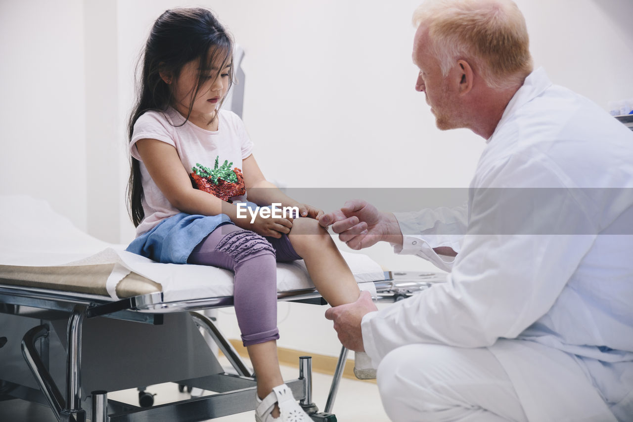 Mature doctor examining girl's knee sitting on bed at hospital