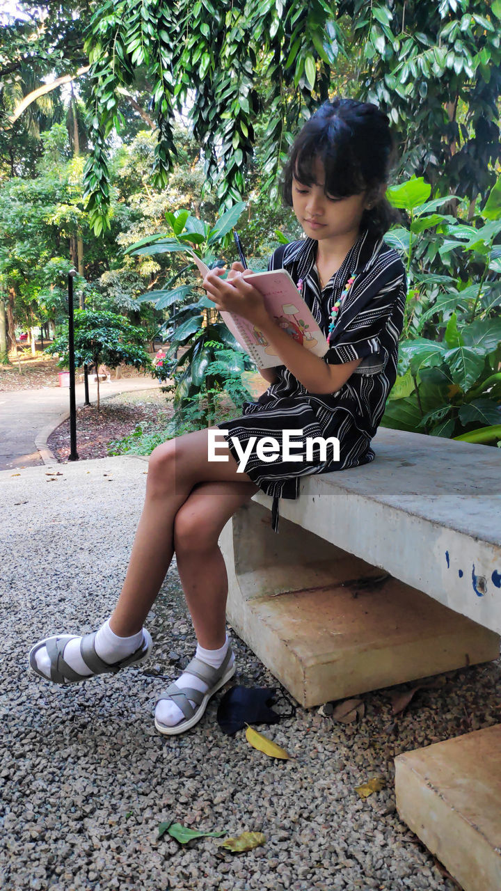 one person, full length, sitting, plant, tree, casual clothing, leisure activity, women, lifestyles, nature, day, looking, person, bench, child, outdoors, young adult, footwear, adult, female, childhood, side view, clothing, looking down, hairstyle, spring, photo shoot, reading, seat, communication, human leg, activity, wireless technology