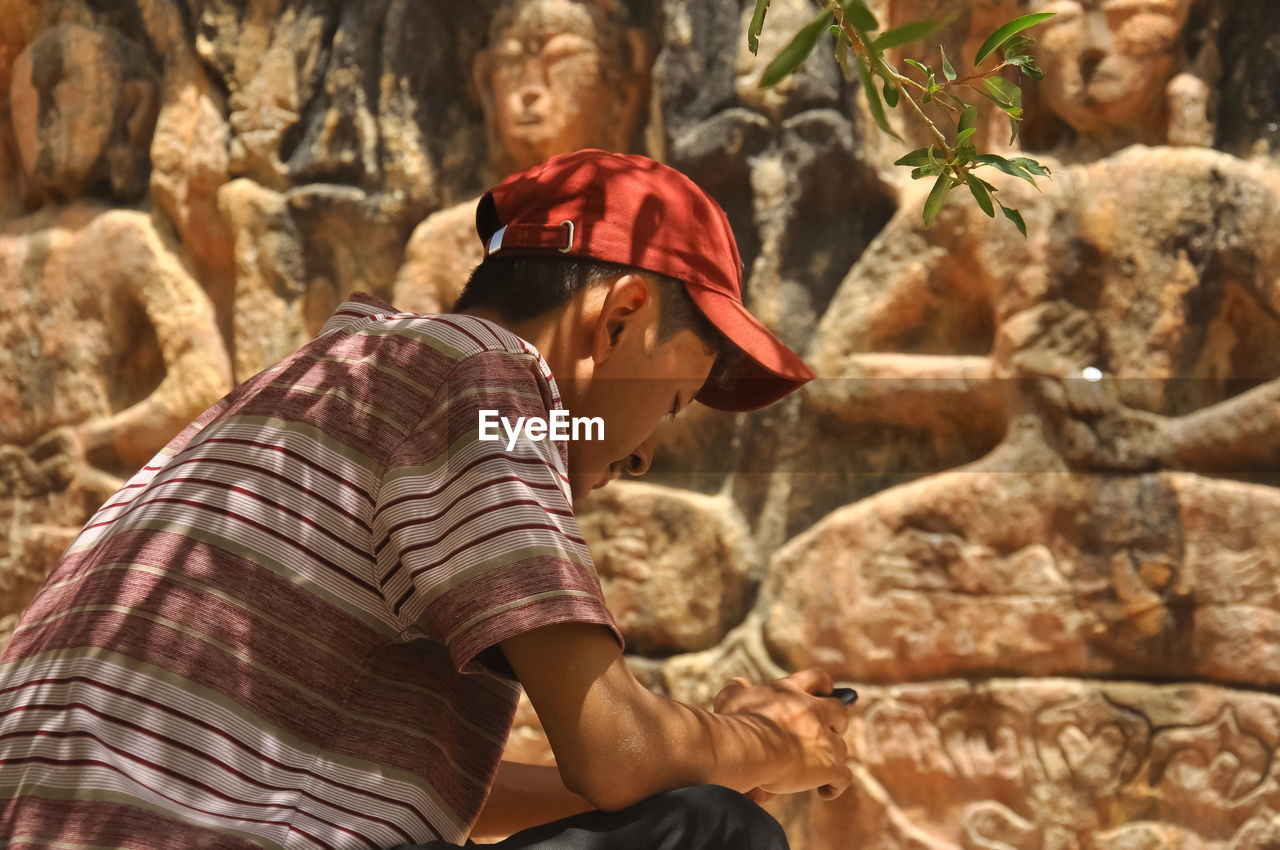 Rear view of a guy using phone while sitting in front of gyalwa ringna 5 dhyani buddha rock statue