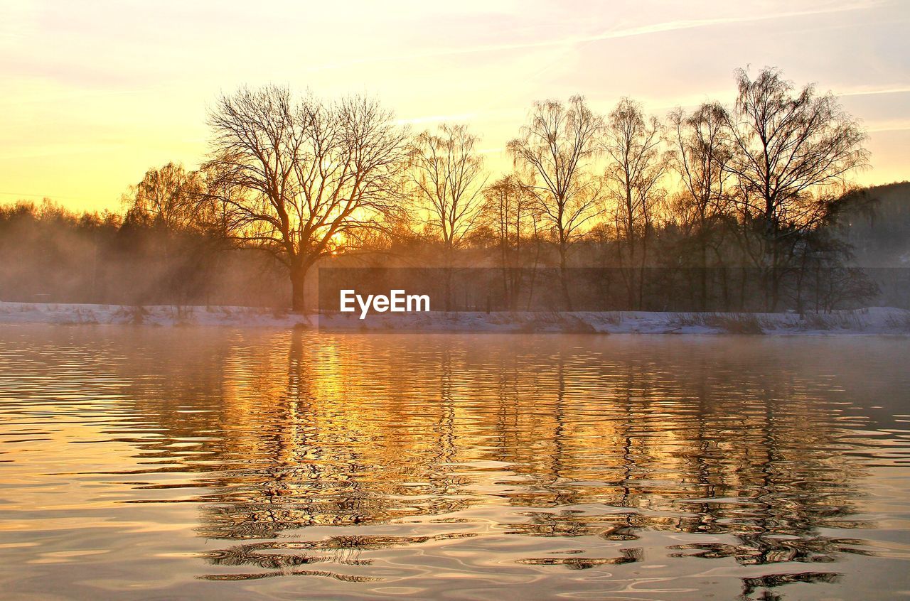 Bare trees by lake against sky during sunset