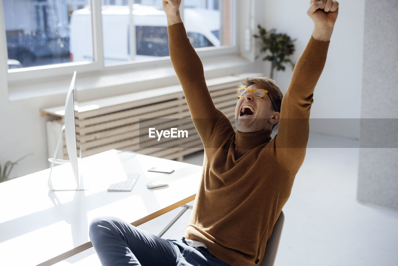 Happy businessman with arms raised shouting sitting at desk in office