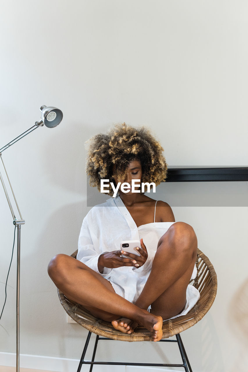 Full body of african american female sitting with crossed legs in wicker armchair and using cellphone