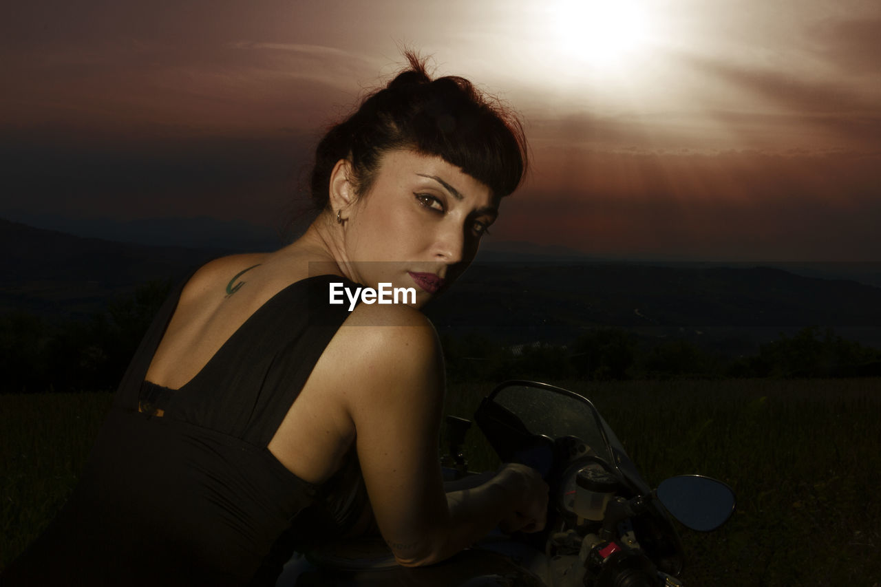Portrait of woman with motorcycle on land against sky during sunset