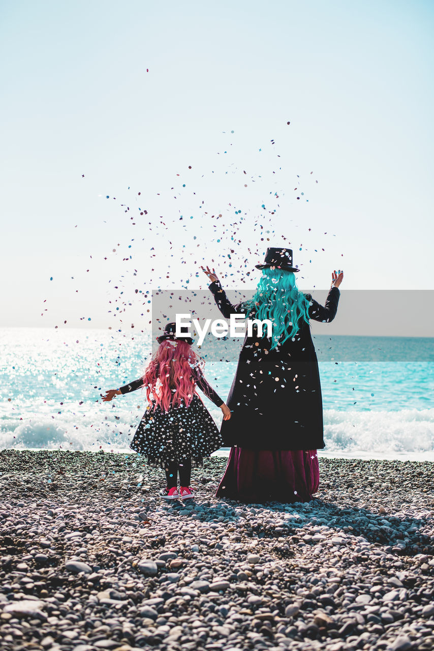Rear view of mother and daughter standing at beach against clear sky and throwing confettis