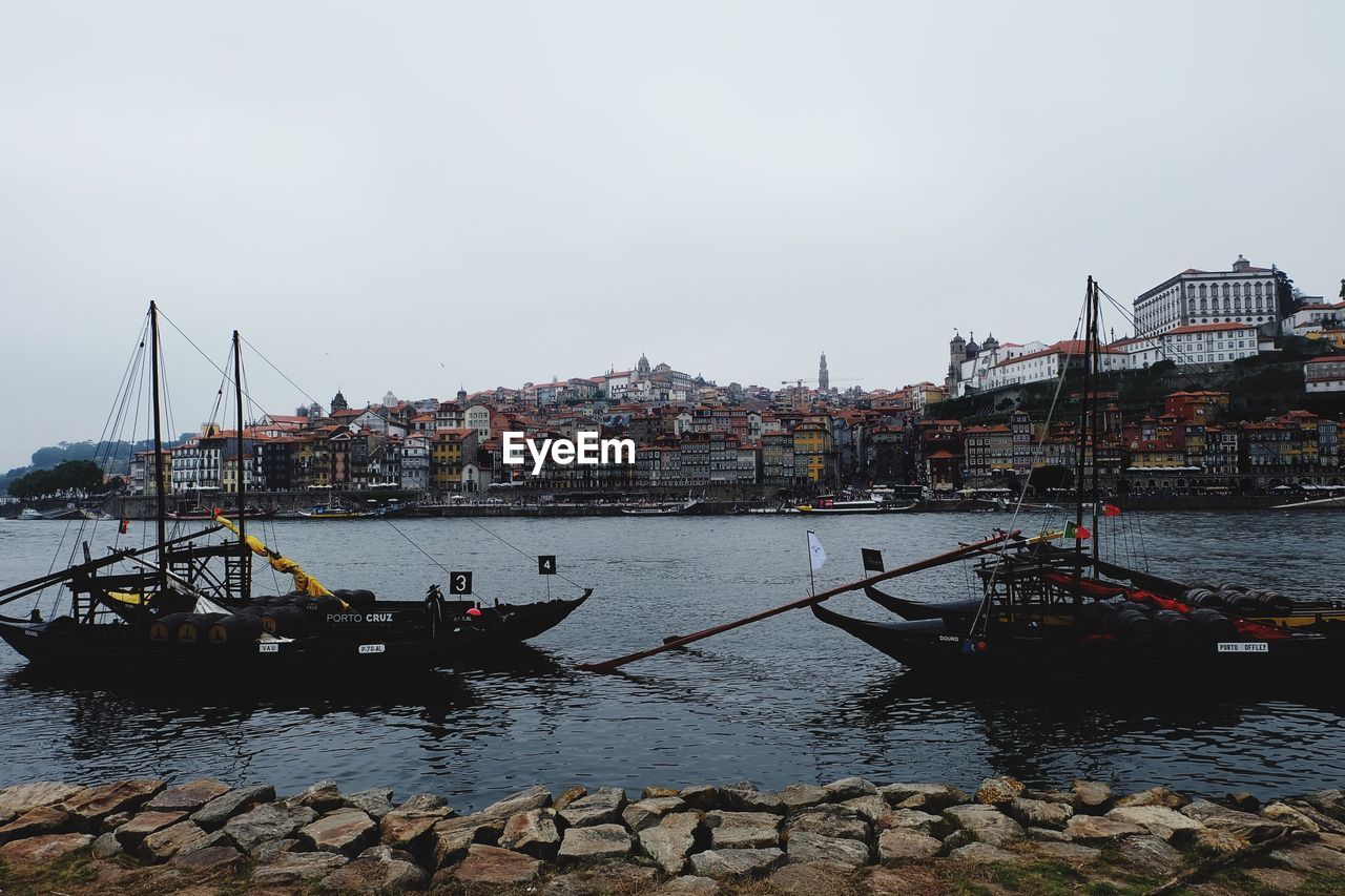 SAILBOATS MOORED IN RIVER BY BUILDINGS AGAINST SKY