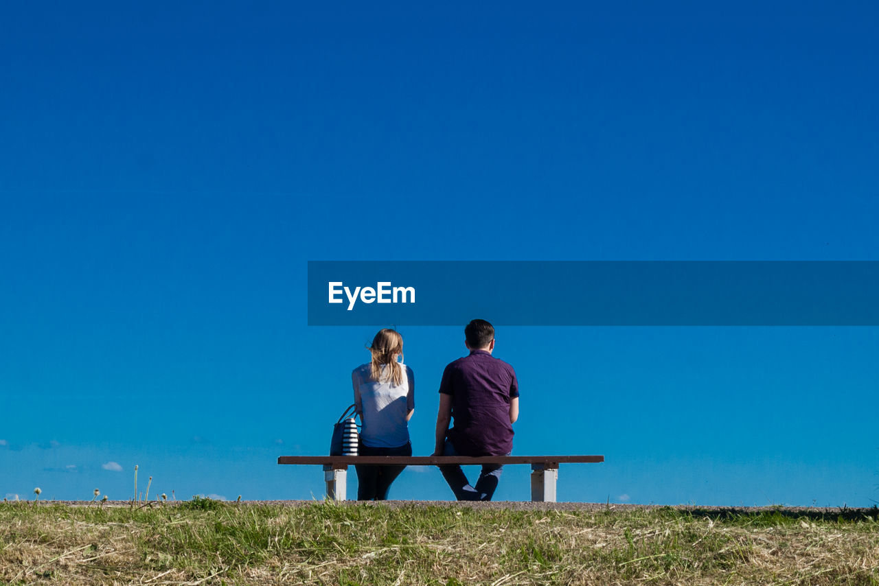 Couple sitting on bench against clear blue sky