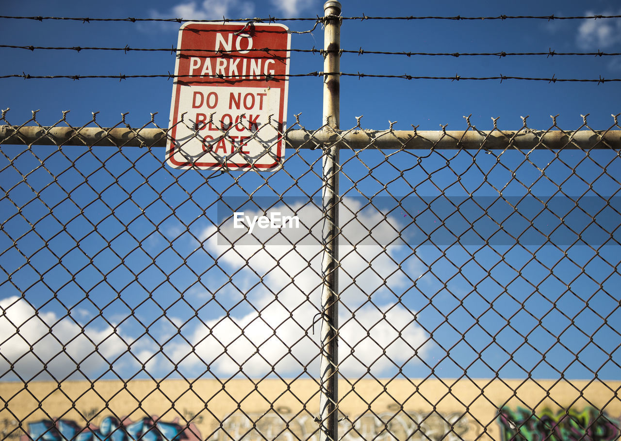 Close-up of information sign hanging chainlink fence against sky