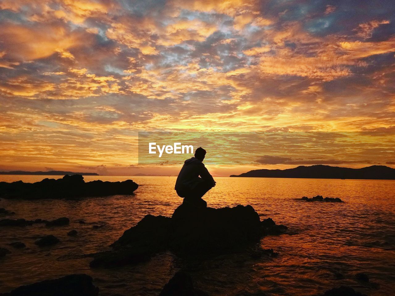 Silhouette man crouching on rock by sea against sky during sunset