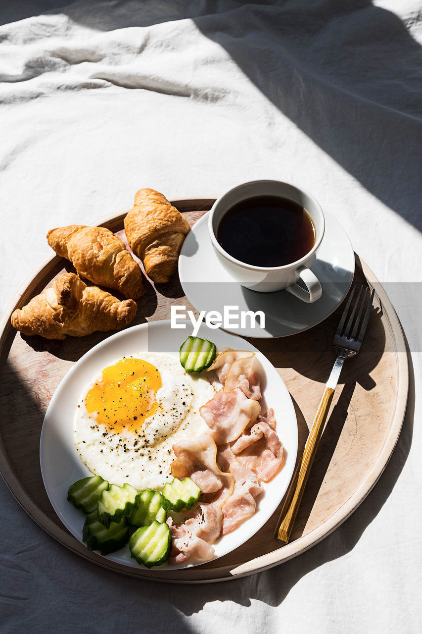 Breakfast in bed coffee cup, fried egg, bacon and croissants on wooden tray. 