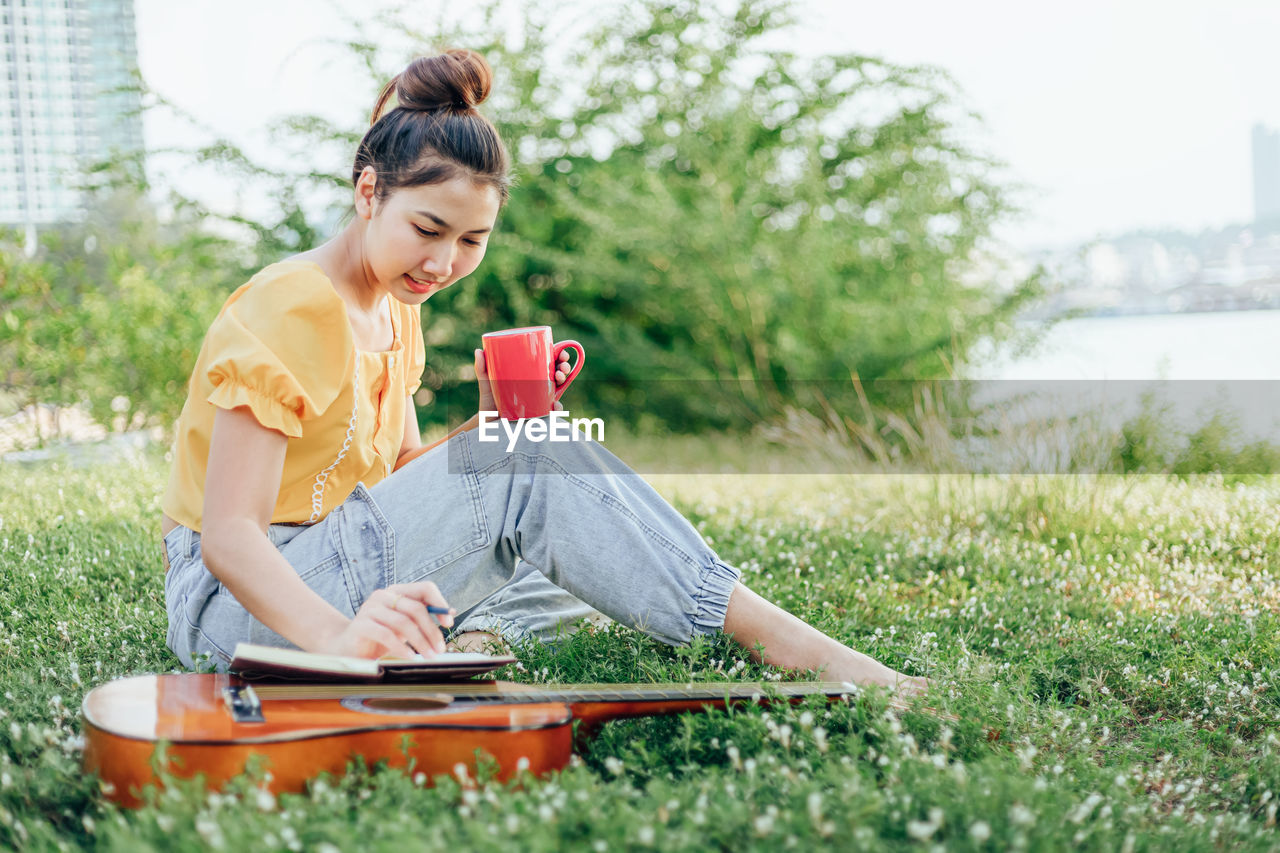 Full length of woman writing on book while having drink at park