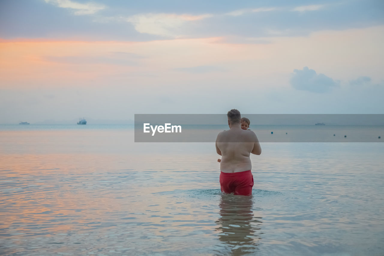 rear view of shirtless man standing at beach against sky during sunset