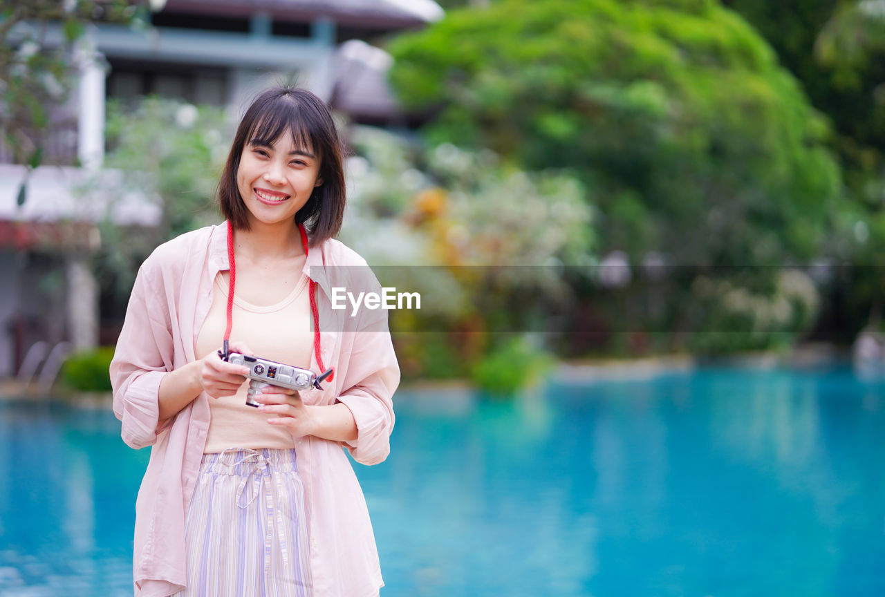 WOMAN HOLDING CAMERA WHILE STANDING AT SWIMMING POOL