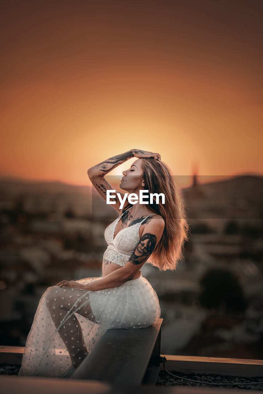 SIDE VIEW OF WOMAN STANDING AGAINST ORANGE SUNSET SKY