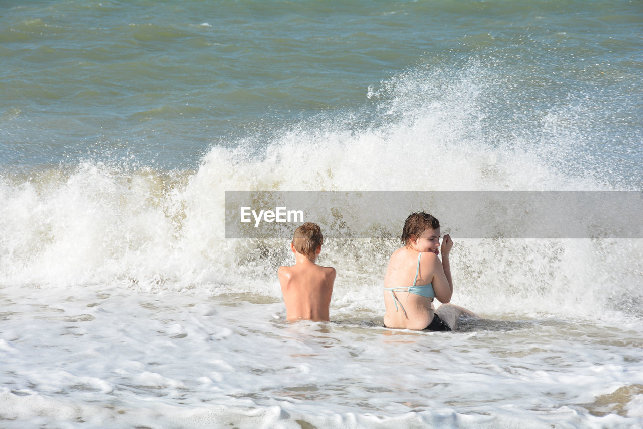 Two children sit on the sandy beach in the middle of the waves by the sea, netherlands