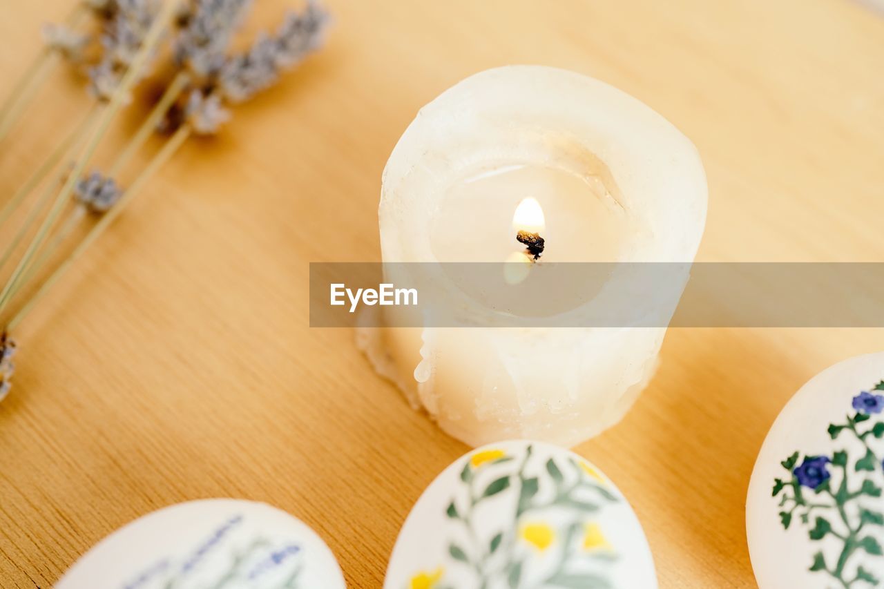 Top view of burning candle surrounded of chicken eggs painted with aquarelle placed on table with lavender flowers for easter