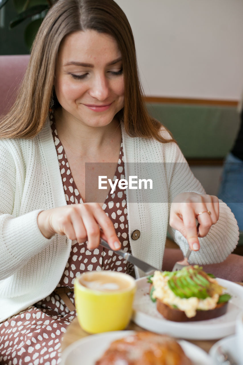 Portrait of young woman sitting on table having brunch in the restaurant, cutting avocado toast