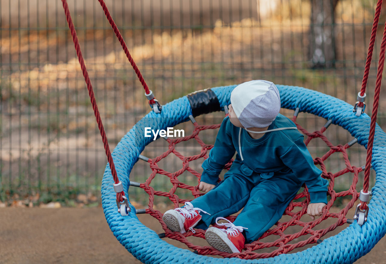 Cute little boy with down syndrome in a funny hat walks in the playground, swinging on a swing