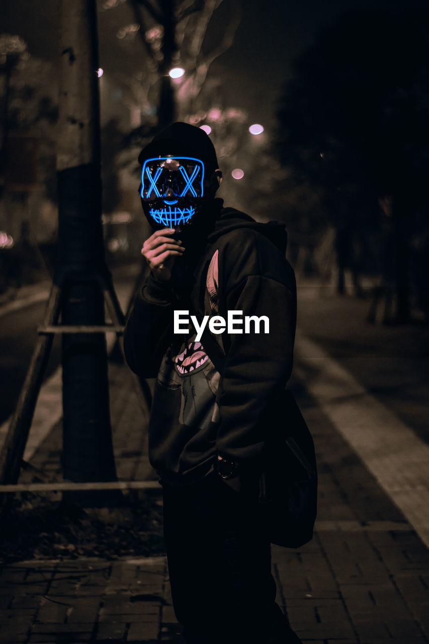 Portrait of the unknown man behind the neon mask on street at night. dark brown tone