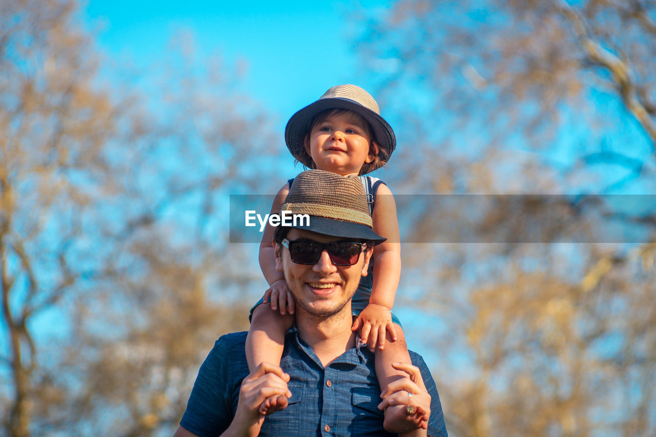 Portrait of happy father carrying son on shoulders against trees