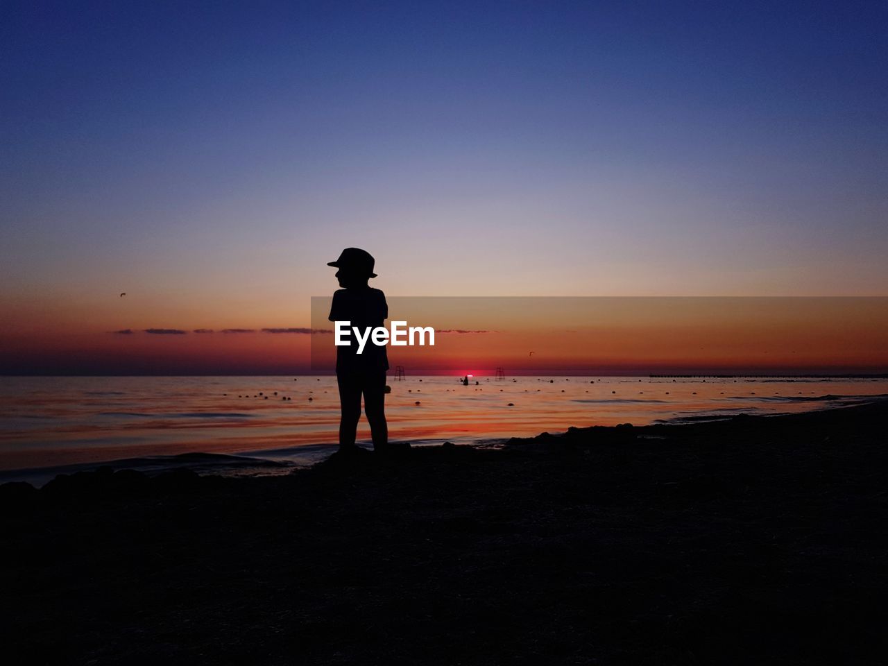 Silhouette boy standing at beach against sky during sunset