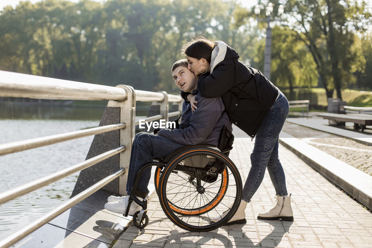 Girlfriend kissing boyfriend sitting in wheelchair at embankment
