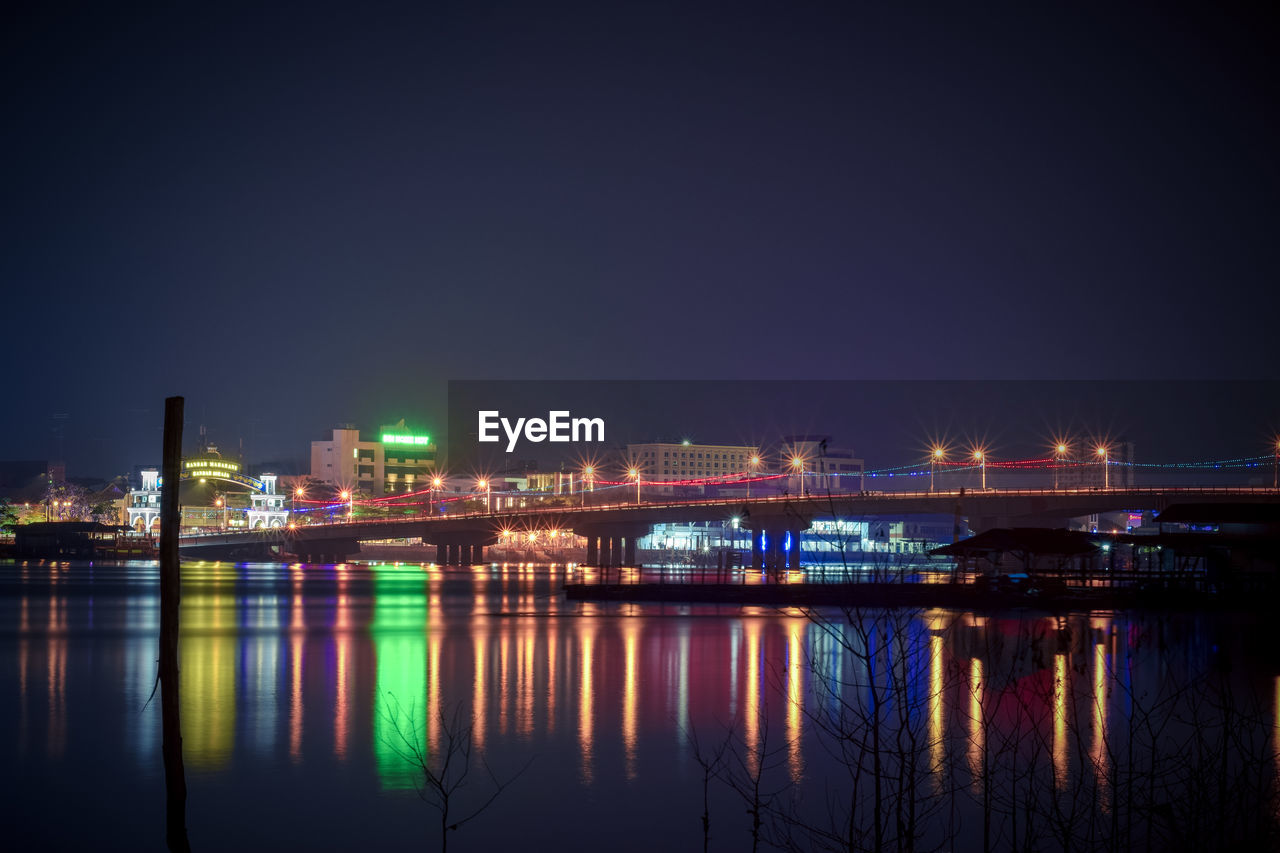 Illuminated buildings by river against sky at night
