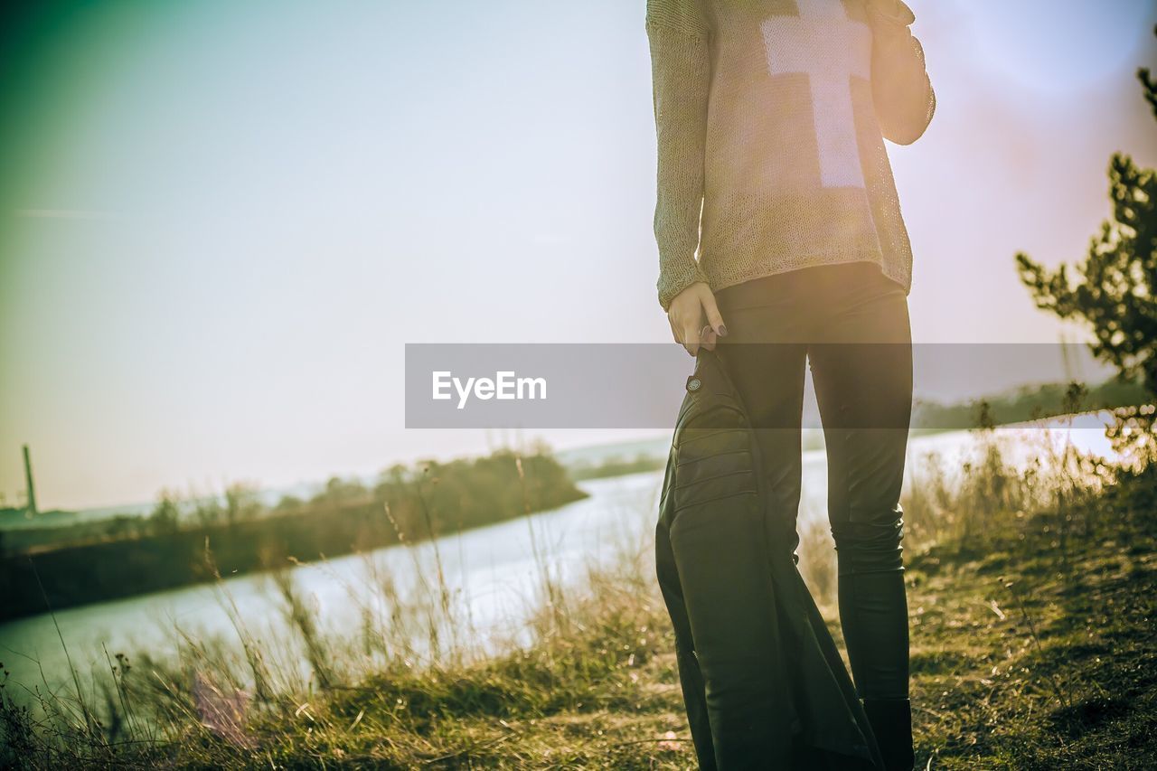 Low section of woman holding motorcycle jacket standing on grass by lake