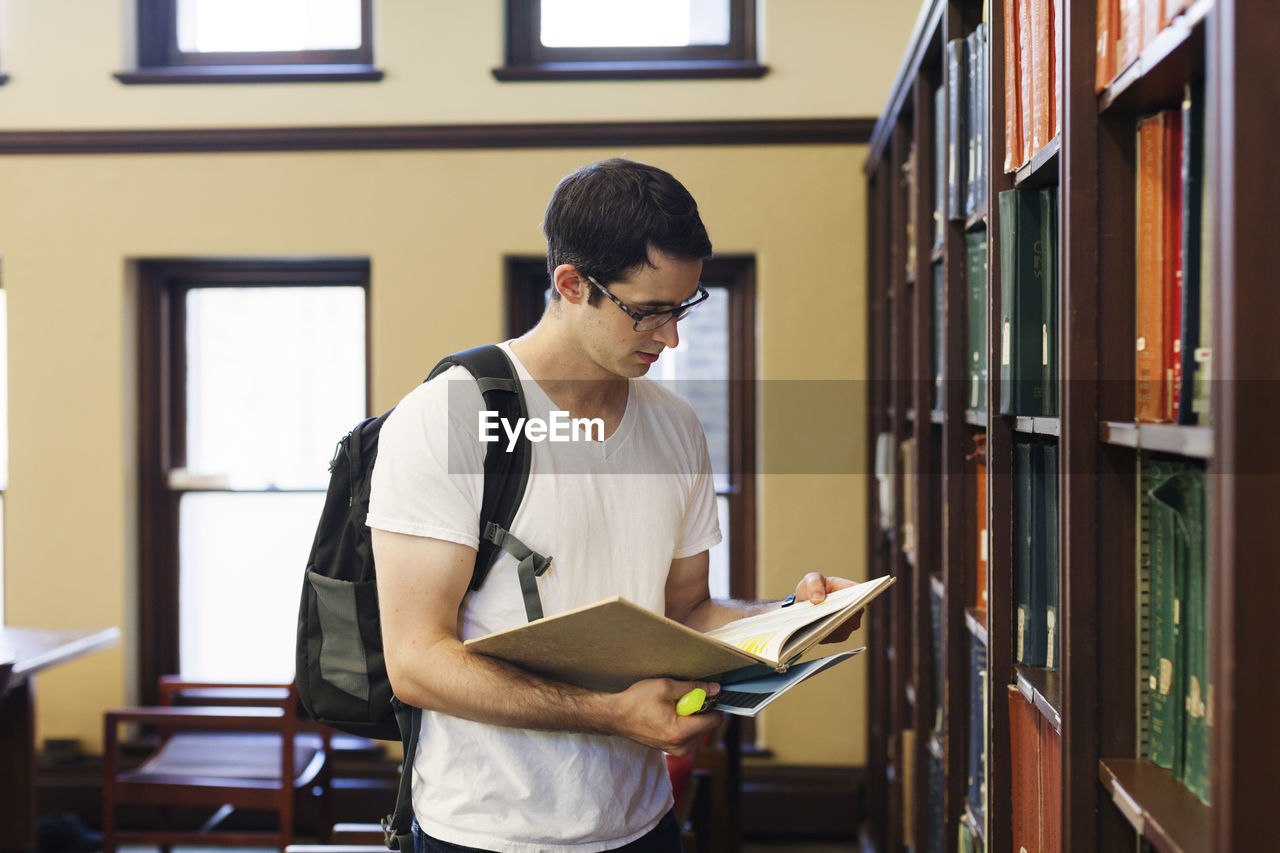Confident man with backpack reading book while standing by shelf in library