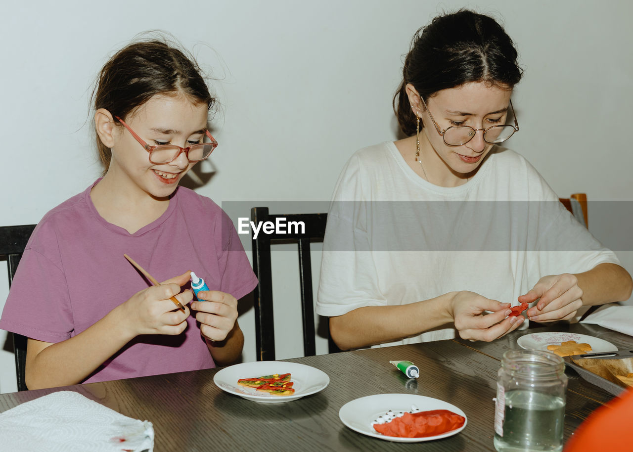 Two girl is coloring cookies while sitting at the table.