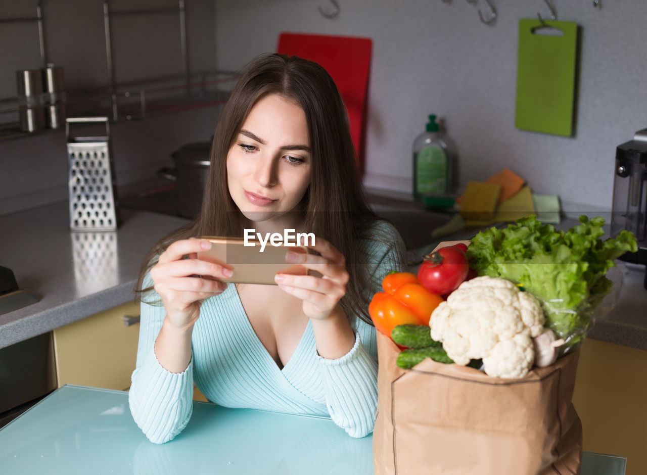 Young woman using mobile phone by vegetables in kitchen at home