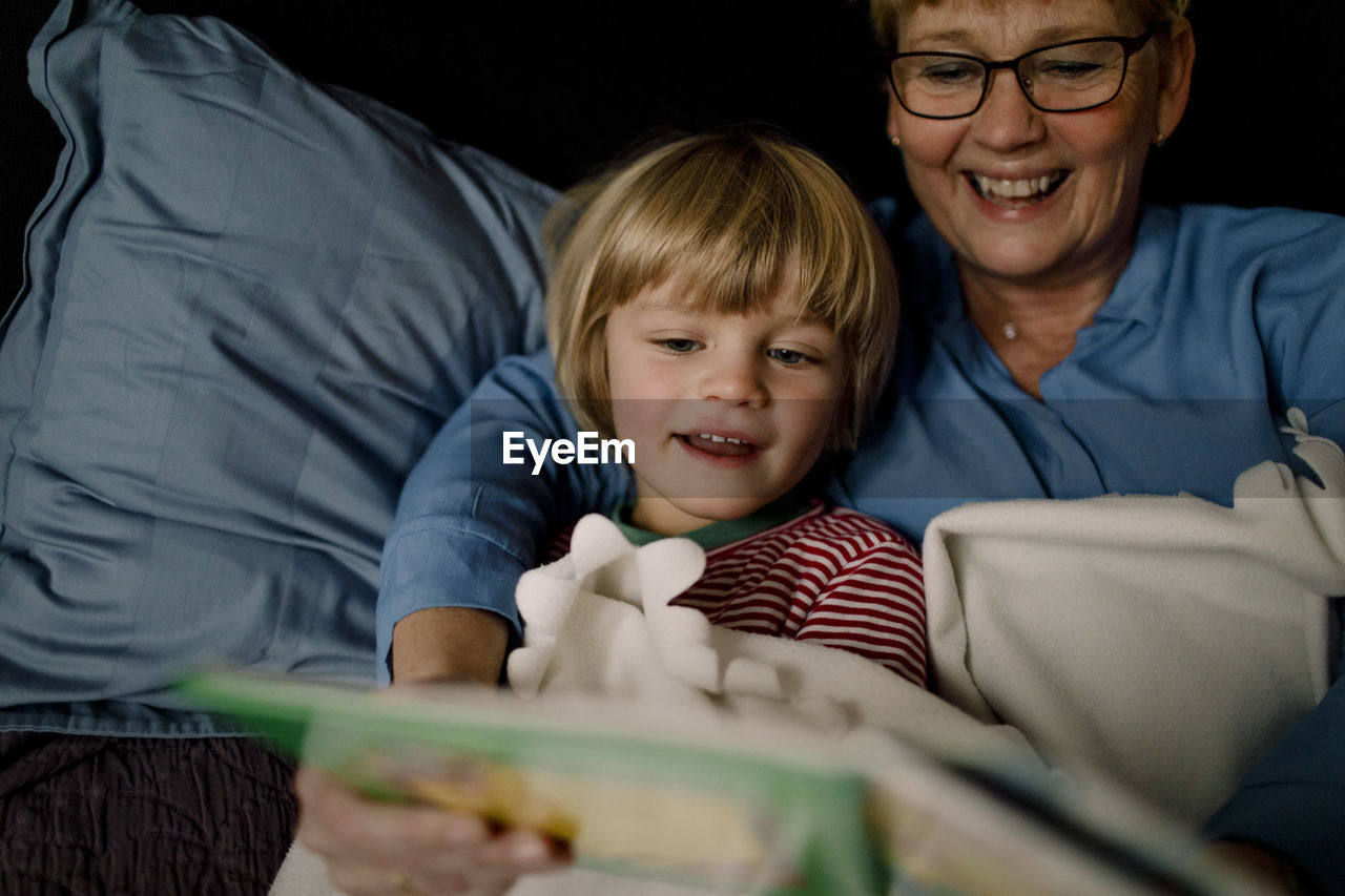 Smiling grandmother reading story book for grandson on bed at home