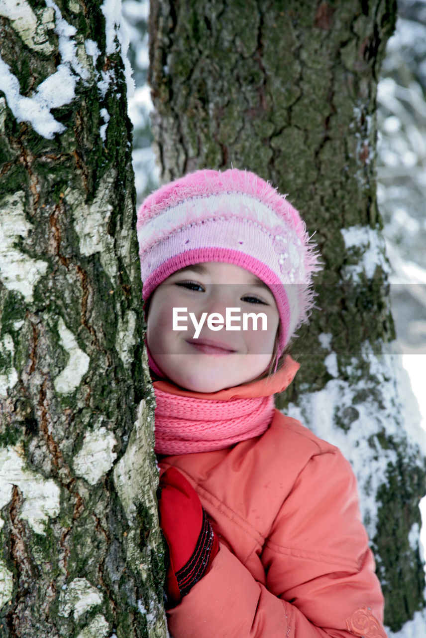 Portrait of happy girl hiding behind tree trunk in winter