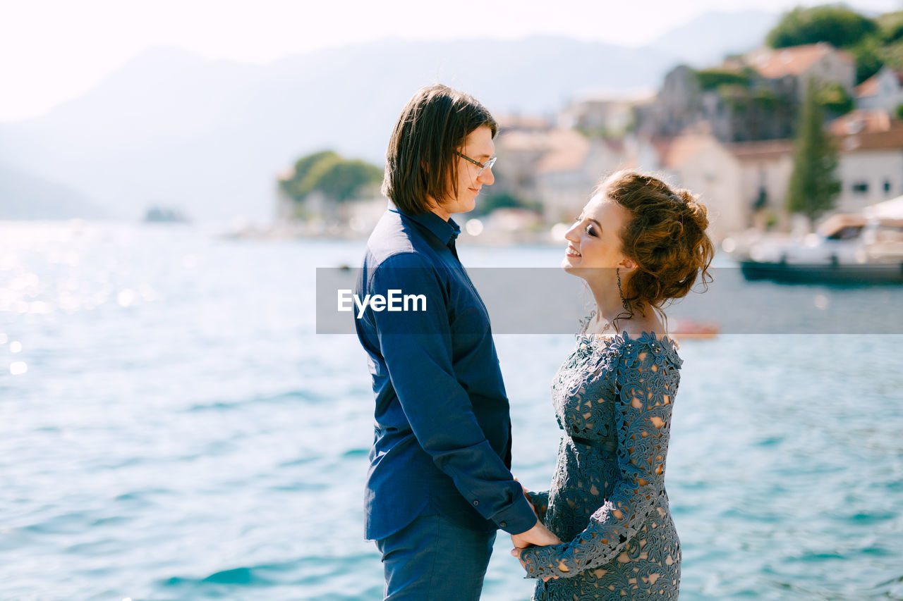 Side view of husband wife holding hands standing against sea