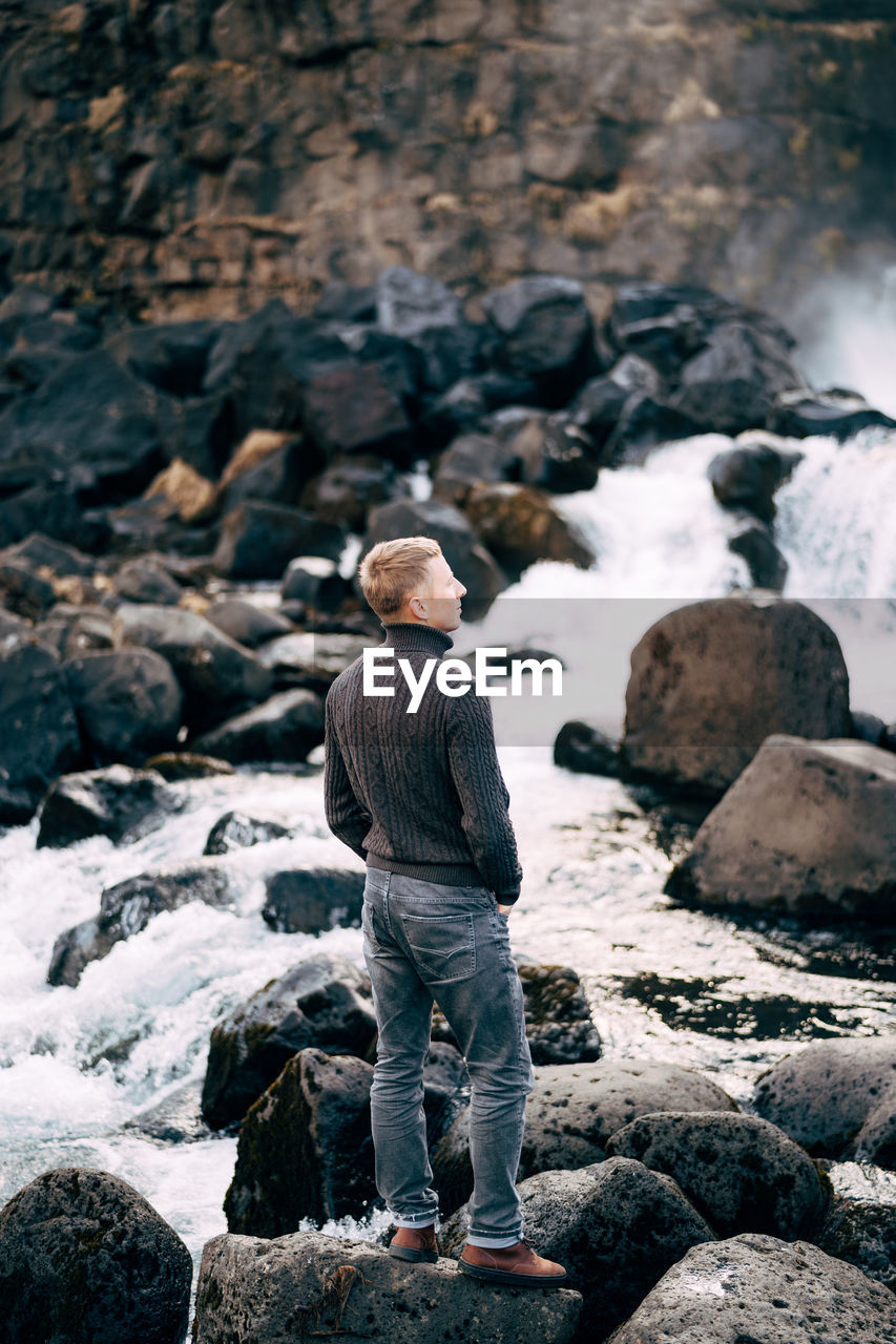 REAR VIEW OF MAN STANDING ON ROCK IN WATER