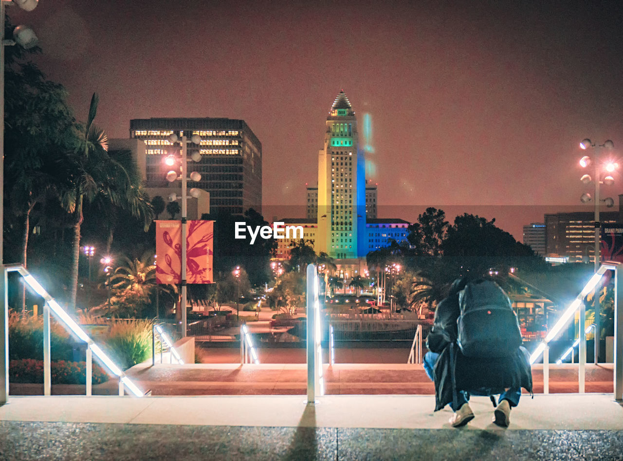 Rear view of person crouching in illuminated garden by buildings at night