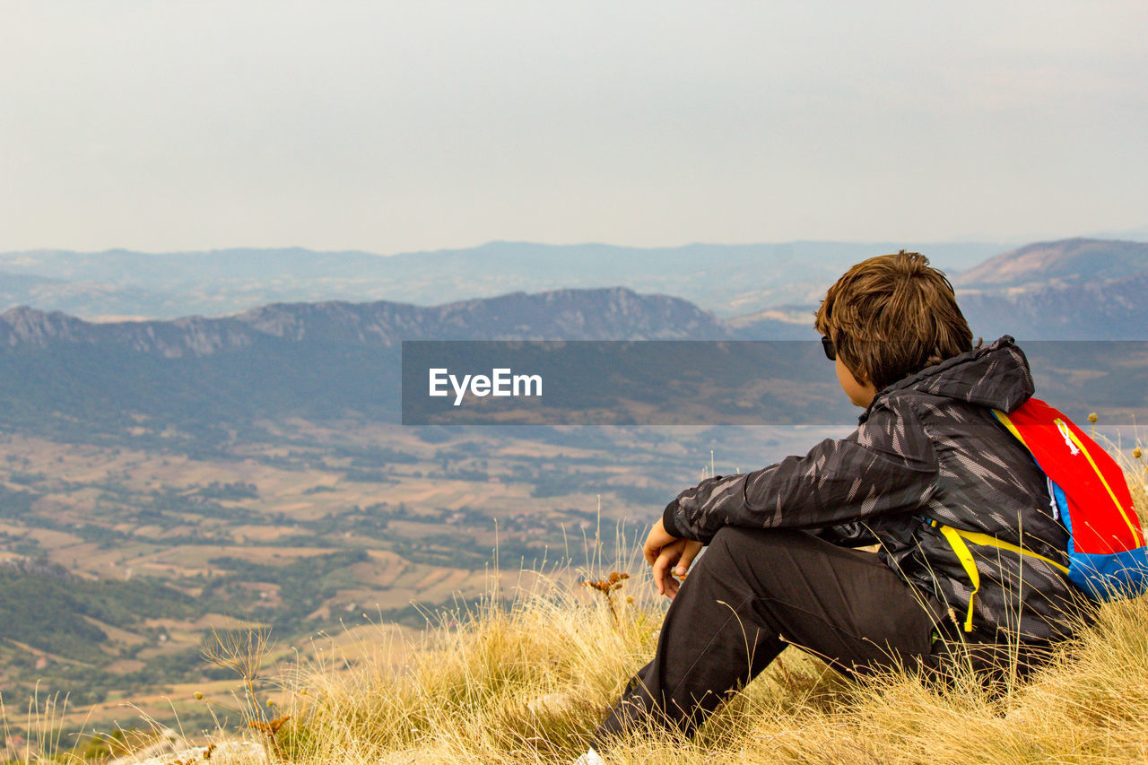 Side view of boy sitting on grassy mountain against sky