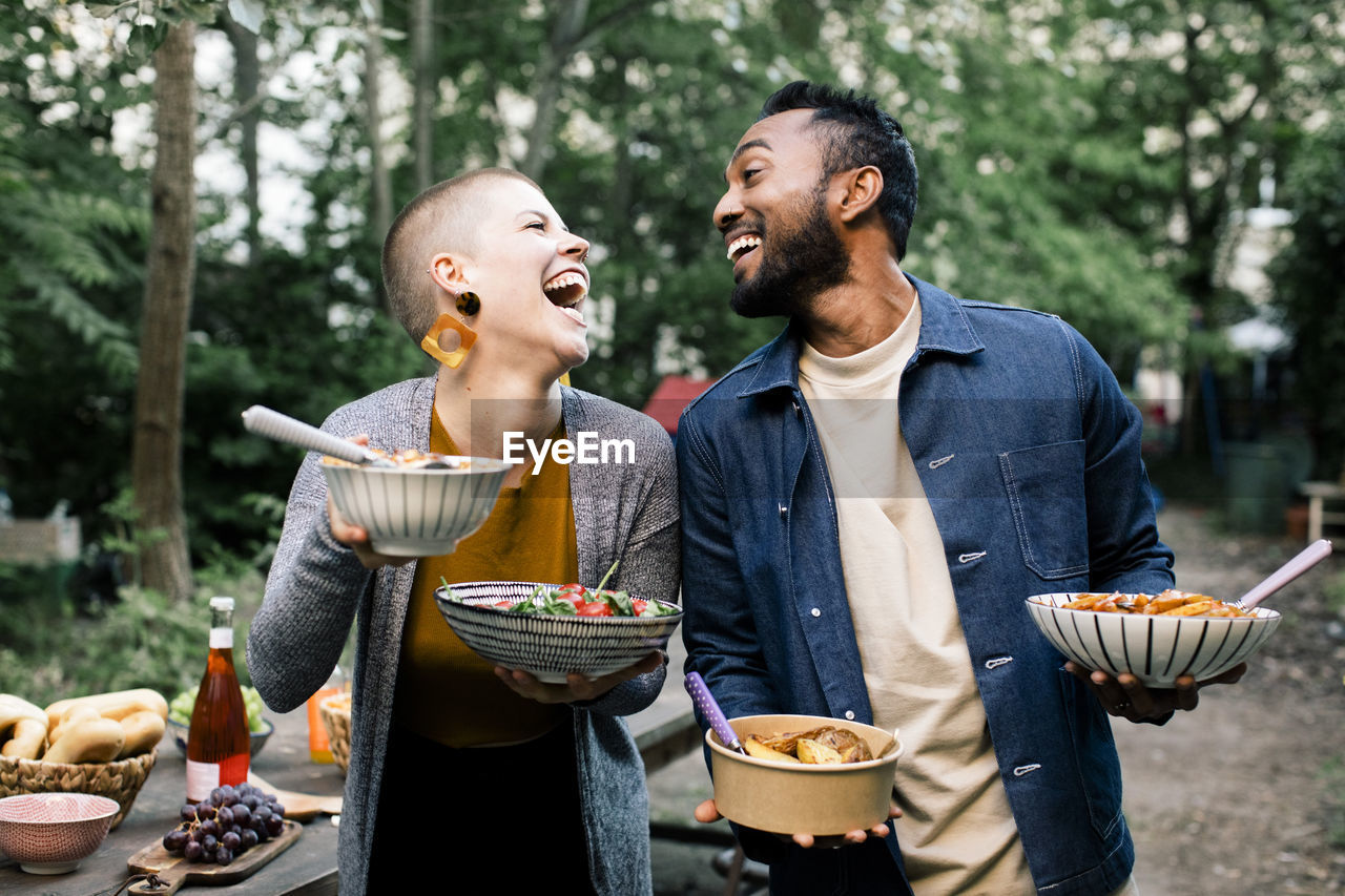 Multiracial male and female friends laughing and looking at each other while holding food bowls