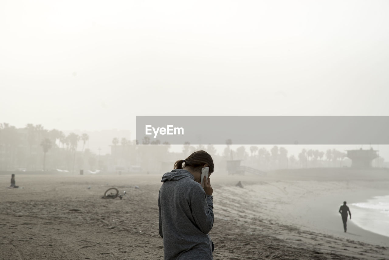 Woman talking on phone while standing at beach against clear sky