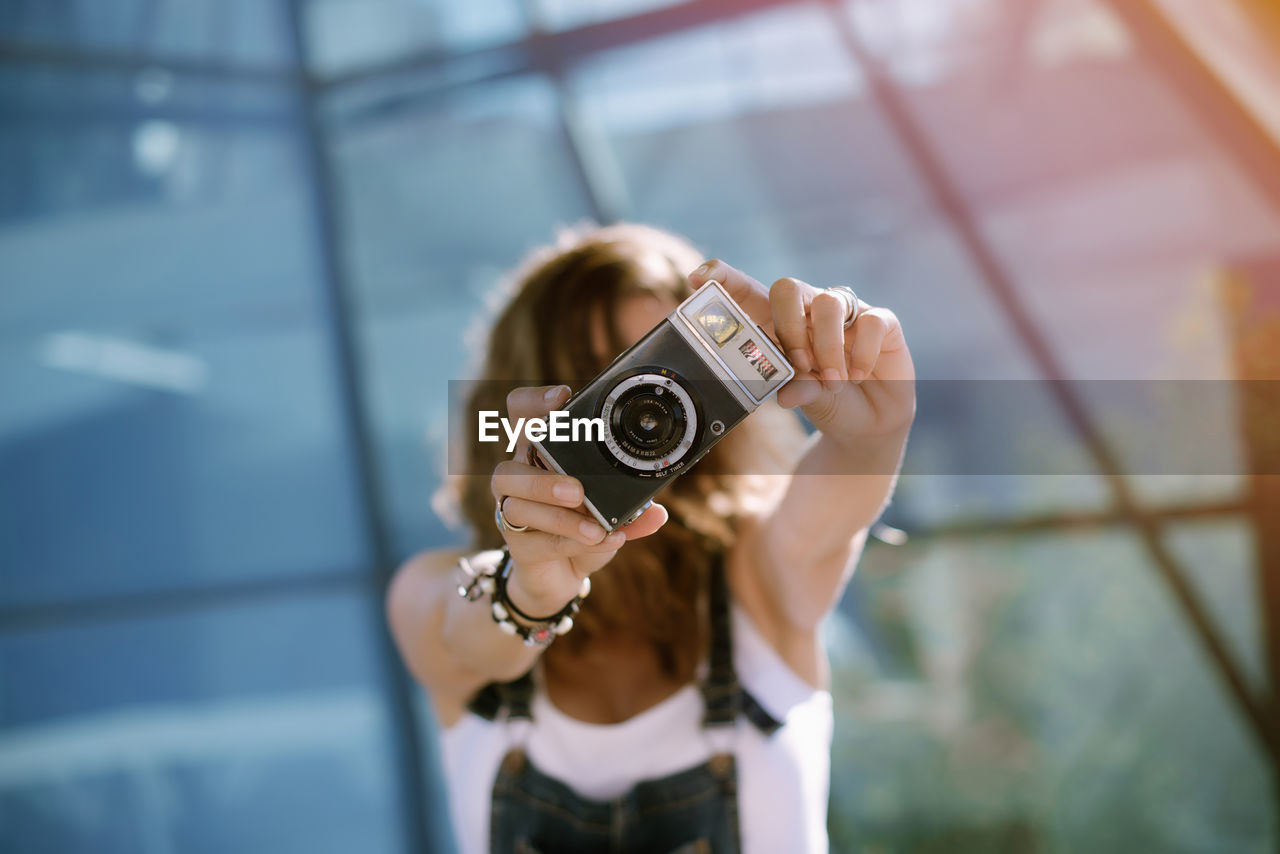 From above of young unrecognizable female holding photo camera in outstretched hands and taking picture while standing against glass wall