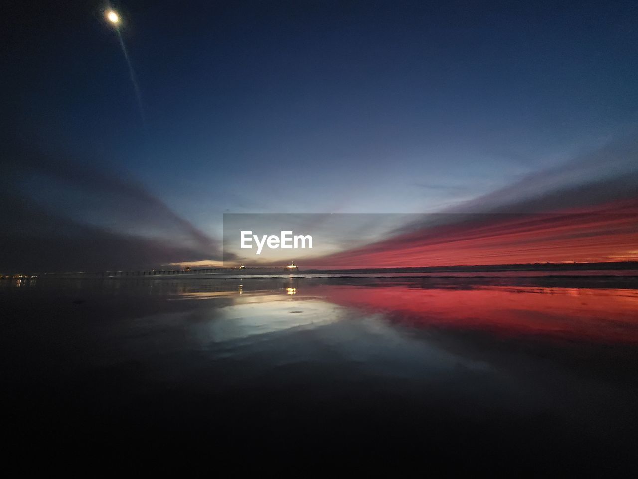 ILLUMINATED ROAD BY SEA AGAINST SKY AT SUNSET