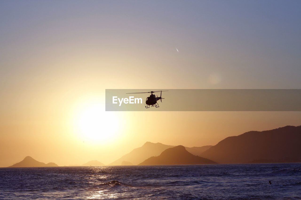 Low angle view of helicopter over sea in front of mountains during sunset