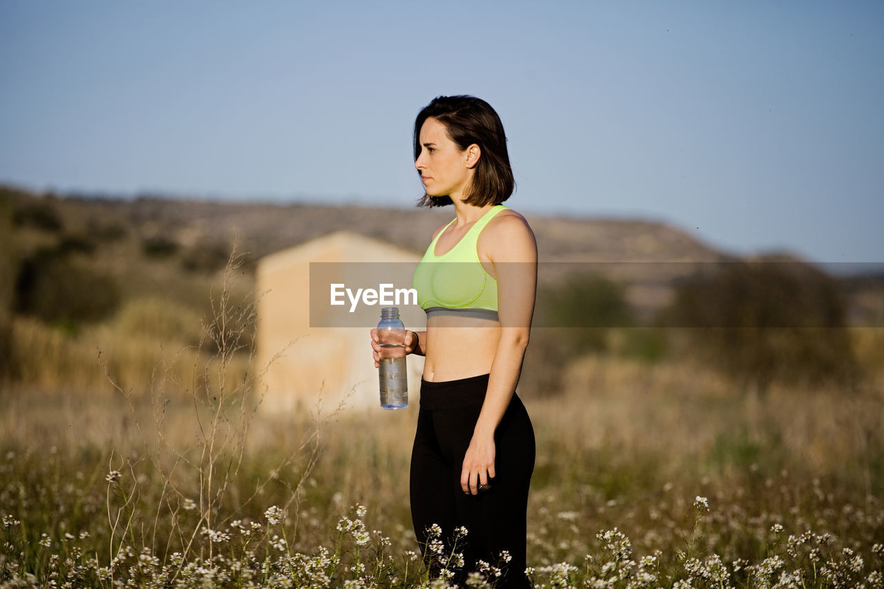 Side view of woman with water bottle standing amidst plants against clear sky