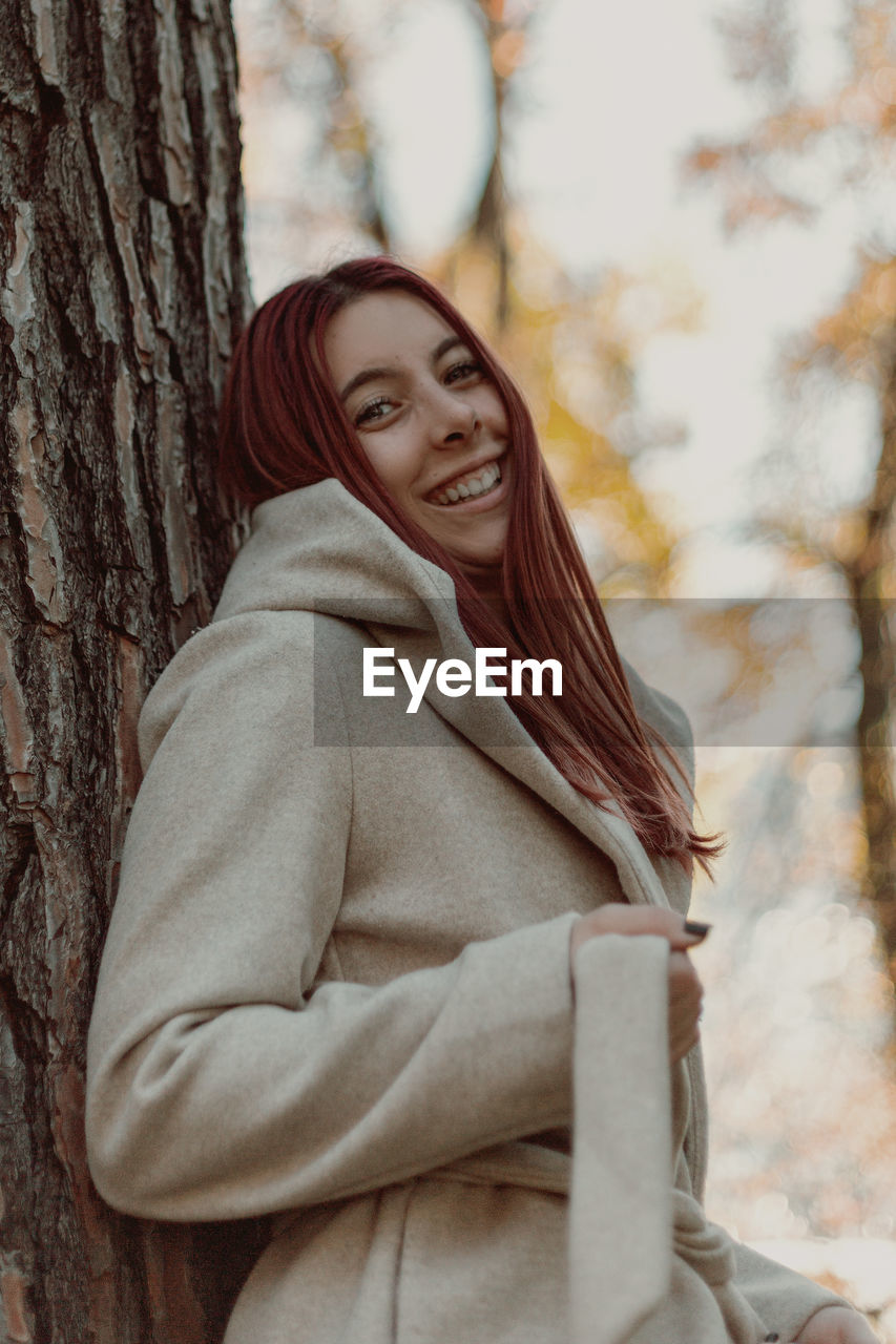 Portrait of smiling young woman standing against tree trunk