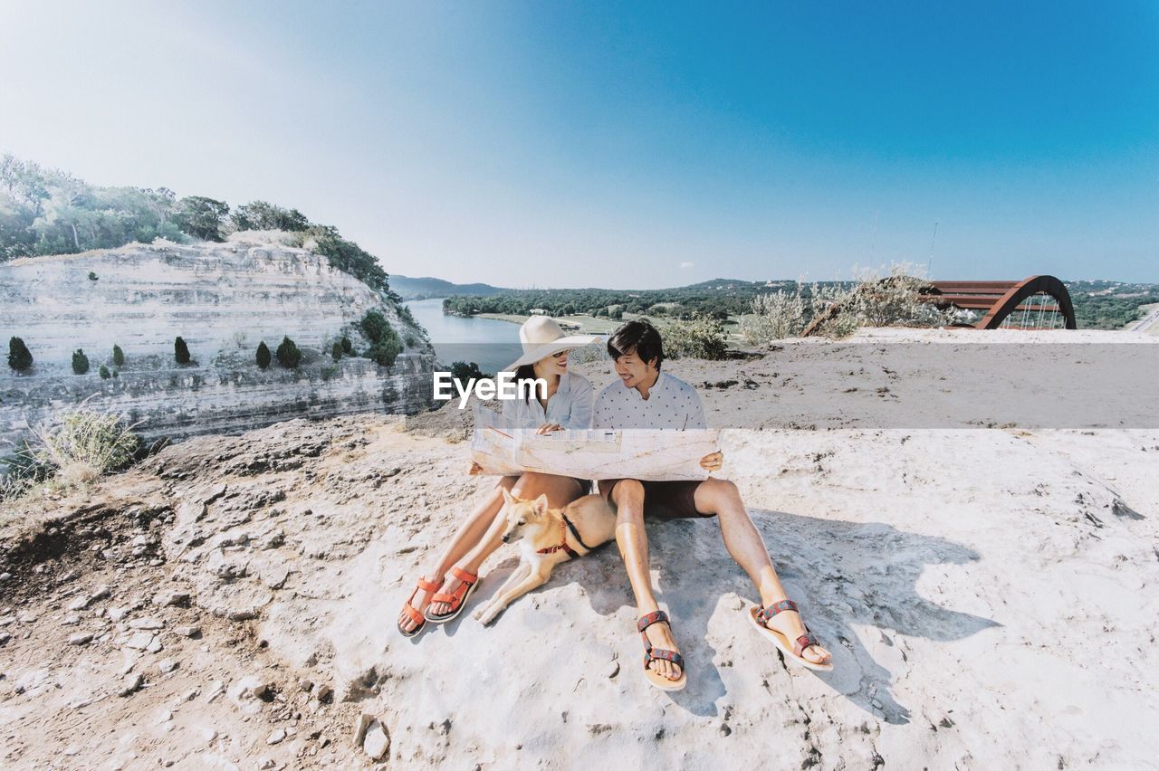 Couple reading map while sitting with dog on rock against clear sky